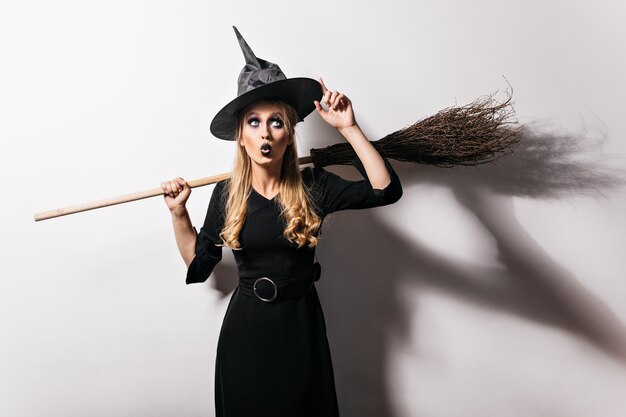 Bruja rubia asombrada tocando su sombrero mágico. Atractiva chica vampiro preparándose para el carnaval de halloween.