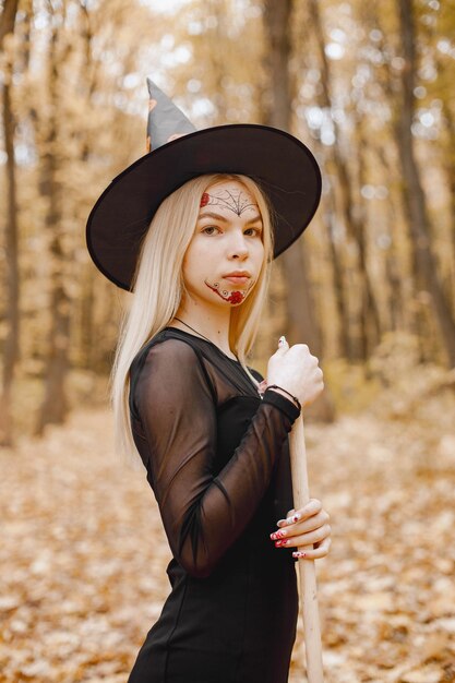 Bruja rubia joven en el bosque en Halloween. Chica con vestido negro y sombrero de cono. Bruja sosteniendo una escoba.