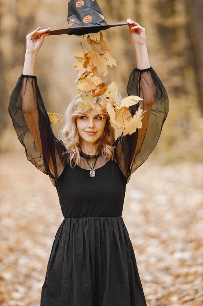 Bruja rubia joven en el bosque en Halloween. Chica con vestido negro y sombrero de cono. Bruja sosteniendo un sombrero y tirando hojas secas.