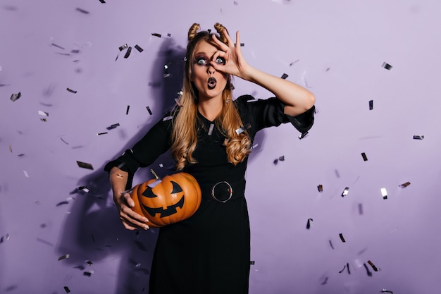 Bruja rubia sorprendida de pie bajo confeti. Conmocionada joven posando en la pared púrpura con calabaza de halloween.