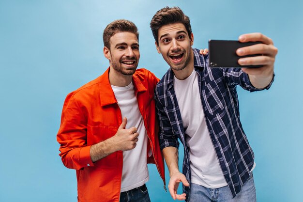 Brunet hombre barbudo en jeans y camisa a cuadros se toma selfie con un amigo Cool guy en chaqueta naranja muestra el pulgar hacia arriba sobre fondo azul