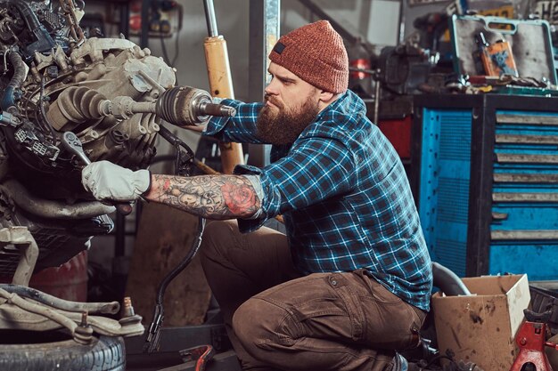 Brutal especialista en mecánica barbudo tatuado repara el motor del coche que se levanta en el ascensor hidráulico en el garaje. Estación de servicio.