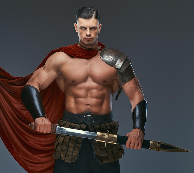 Brutal guerrero de la antigua Grecia con un cuerpo musculoso en uniformes de batalla posando sobre un fondo gris.