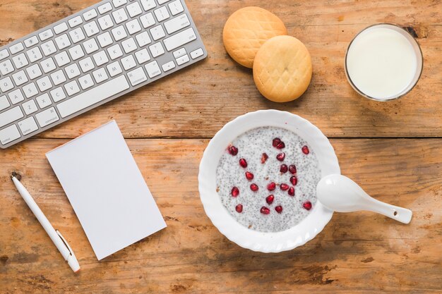 Budín de semilla de chía; galletas; zalamero; Leche; bloc; Pluma y teclado de computadora inalámbrico sobre fondo de madera