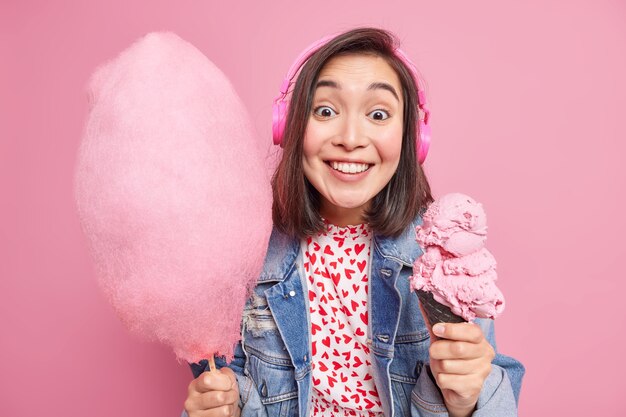 Buen aspecto positivo morena modelo femenino asiático sonríe positivamente vestida con ropa de moda posa con deliciosos postres dulces apetitosos contra la pared rosa. Disfruta de helado y algodón de azúcar.