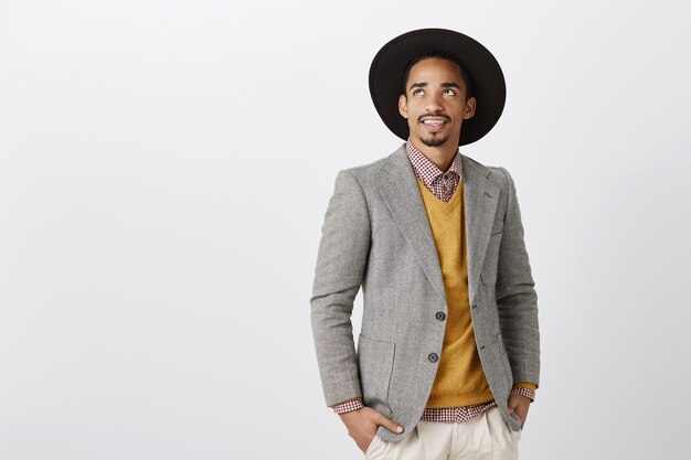 Buen día para ganar miles de millones. Retrato de hombre de negocios rico y guapo con elegante traje formal y sombrero mirando hacia arriba, interesado y entretenido con algo curioso, soñando sobre una pared gris