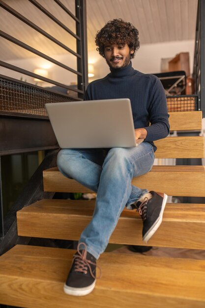 Buen humor. Sonriente joven adulto indio con suéter azul y jeans interesado en trabajar en una laptop sentada en las escaleras en una habitación iluminada