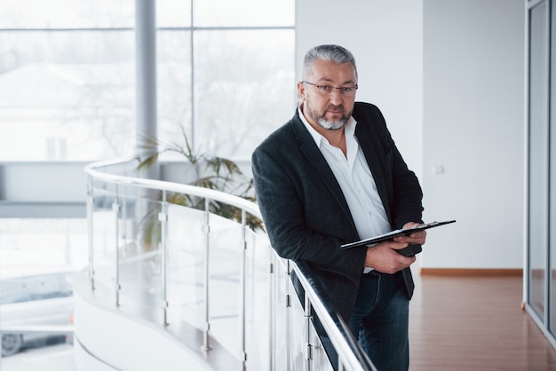 Buen retrato Foto del empresario senior en la espaciosa habitación con plantas detrás. Sosteniendo y leyendo documentos