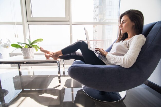 Buen tiempo en casa. Hermosa mujer sonriente joven que trabaja en la computadora portátil mientras está sentado en una silla grande y cómoda en casa