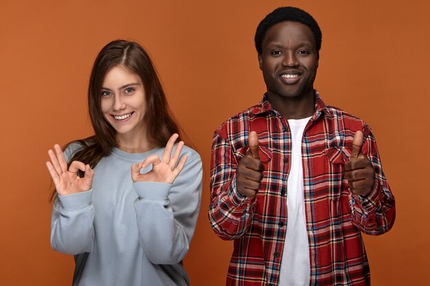 Buen trabajo. Bien hecho. Feliz mujer caucásica joven positiva vestida con una sudadera de gran tamaño haciendo un gesto de ok y un hombre afroamericano alegre y guapo mostrando los pulgares hacia arriba como símbolo de aprobación y me gusta