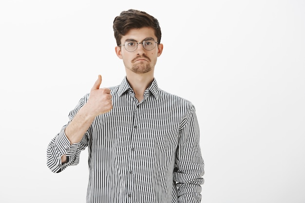 Buen trabajo, hombre. Estudiante masculino atractivo e impresionado complacido con gafas, mostrando los pulgares hacia arriba y chupándose los labios, dando aprobación y gustando la idea o proyecto de un amigo, compartiendo su opinión sobre la pared gris