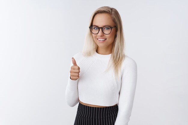 Buen trabajo, orgulloso de ti. Mujer elegante y coqueta atractiva complacida y encantada con gafas y un suéter blanco flaco mostrando los pulgares para arriba gesto y sonriendo impresionado contra el fondo blanco.