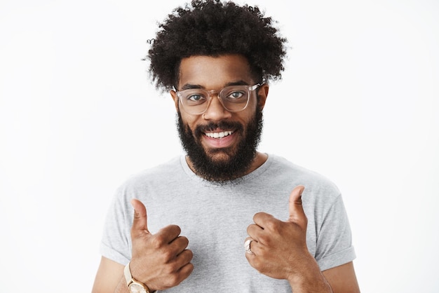 Buen trabajo, orgulloso de ti. Retrato de emprendedor masculino satisfecho, solidario y amistoso con barba, nariz perforada y peinado afro que muestra los pulgares para arriba gesto y sonriendo complacido en la pared gris.