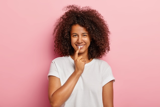 Buena chica divertida con cabello afro muy rizado, sonríe ampliamente, mantiene el dedo índice en los labios, tiene una idea interesante, reflexiona sobre un gran plan, vestida con ropa informal, modelos sobre una pared rosada