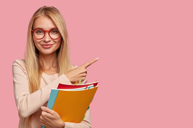 Buena estudiante universitaria rubia posando contra la pared rosa