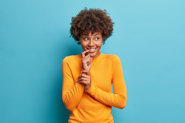 Buena mujer afroamericana alegre sonríe suavemente viste un jersey de manga larga casual parece alegre a un lado posa contra la pared azul