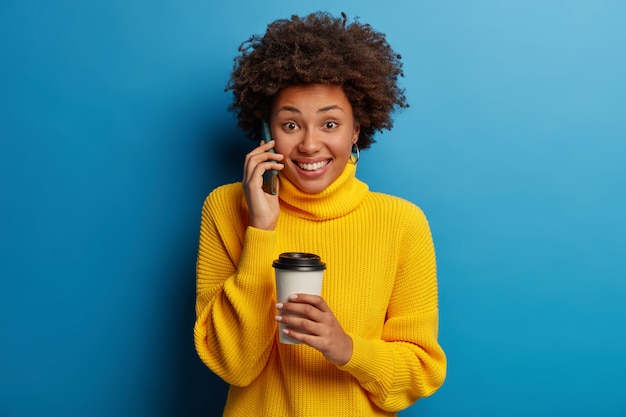 Buena mujer afroamericana positiva tiene conversación telefónica, sostiene el teléfono móvil cerca de la oreja