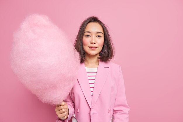 Buena mujer asiática sostiene algodón de azúcar rosa endulzado vestida con ropa formal pasa vacaciones con niños come postre dulce alto en calorías aislado sobre pared rosada. Concepto de estilo de vida