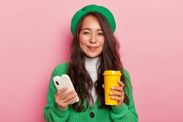 Buena mujer con cabello lacio oscuro, mejillas rojas sostiene un teléfono móvil blanco y una taza de café, disfruta de tiempo libre para navegar por las redes sociales