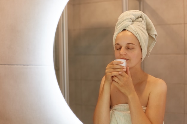 Buena mujer envuelta en una toalla blanca después de tomar una ducha de pie frente al espejo con los ojos cerrados, oler la nueva crema, cuidado de la piel, cosmetología, procedimientos matutinos.