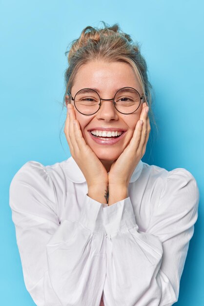 Buena mujer europea encantadora mantiene ambas manos en las mejillas sonríe ampliamente vestida con camisa blanca, lentes transparentes, escucha noticias agradables, posa sobre fondo azul, se ve linda en la cámara