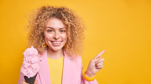 Buena mujer joven con cabello rizado y tupido sonríe agradablemente indica que muestra el lugar para su anuncio contra la pared amarilla sostiene un delicioso helado congelado en gofre