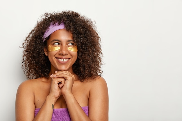 Buena mujer joven de pelo rizado con expresión facial satisfecha, mira a un lado, tiene belleza natural, usa diadema en la cabeza, hace un tratamiento de rejuvenecimiento, se aplica parches de hidrogel debajo de los ojos