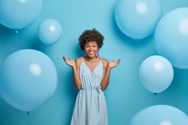 Buena mujer de pelo rizado extiende las palmas de las manos, sonríe sinceramente, disfruta de la fiesta de verano, viste un vestido azul, se opone a los globos de aire festivos, tiene un estado de ánimo feliz, aislado. Feminidad, estilo, concepto de moda.