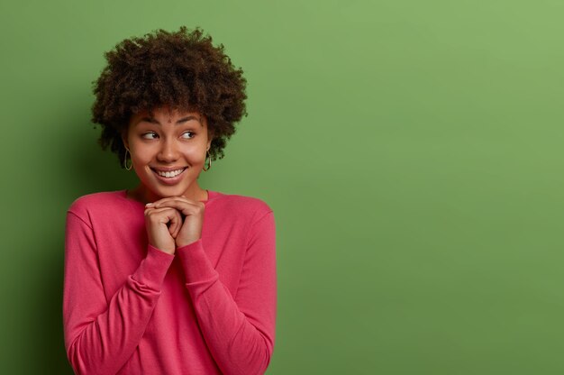 Buena mujer rizada étnica complacida tiene buen humor y expresa emociones positivas, mantiene las manos debajo de la barbilla, mira a un lado, tiene una sonrisa dentuda, vestida con un jersey rosado, aislado en una pared verde