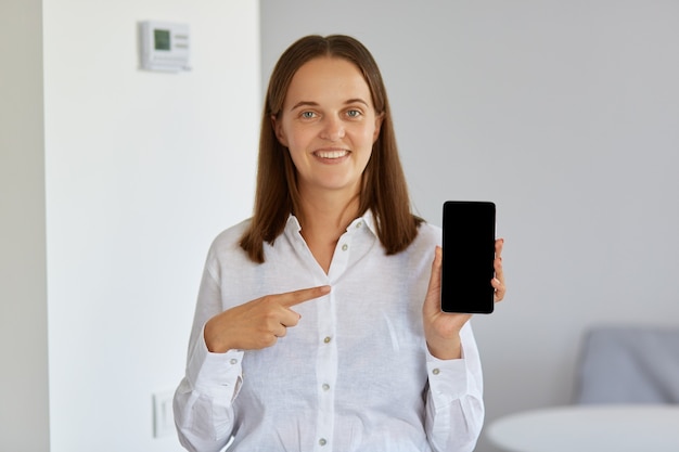 Buena mujer vestida con camisa blanca sosteniendo el teléfono inteligente en las manos y apuntando a su pantalla en blanco con el dedo índice, mirando a la cámara con una sonrisa encantadora.