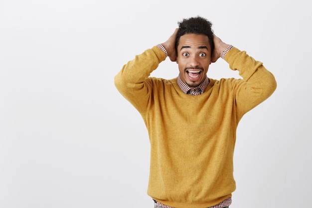 Las buenas noticias inesperadas hacen que el día sea brillante. Chico africano guapo sorprendido en suéter amarillo casual tomados de la mano en la cabeza por la incredulidad, siendo feliz después de un evento positivo, de pie sobre una pared gris