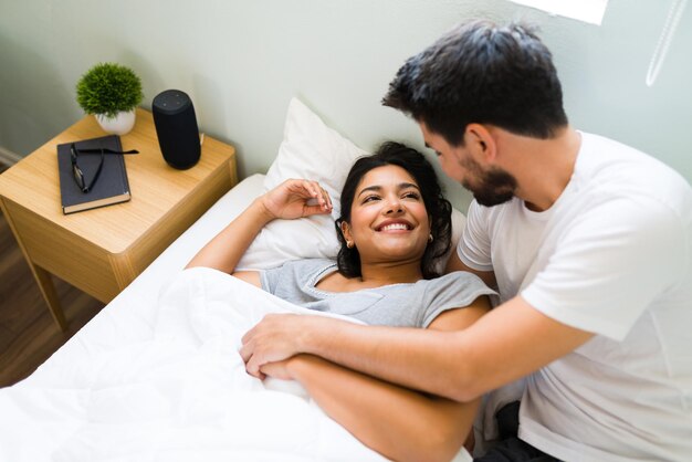 ¡Buenos días cariño! Novio hispano sonriendo a su novia después de despertarla. Pareja feliz durmiendo juntos en una cama cómoda