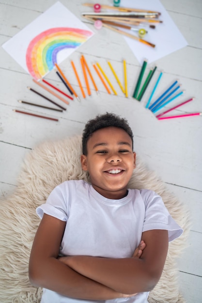 Buenos momentos. Vista superior de un niño en edad escolar sonriente de piel oscura con camiseta blanca tirado en el suelo con las manos dobladas y cerca de lápices de colores, pinceles y dibujos en una habitación luminosa