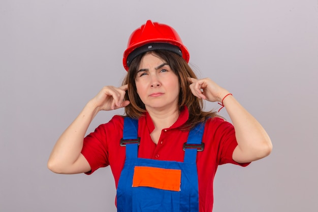 Builder mujer vestida con uniforme de construcción y casco de seguridad cubriendo las orejas con los dedos con expresión molesta por el ruido sobre la pared blanca aislada