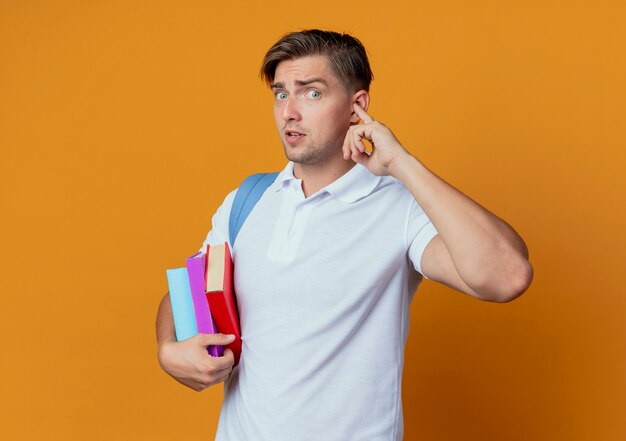 Buscando joven apuesto estudiante masculino con bolsa trasera sosteniendo libros y oído cerrado con el dedo