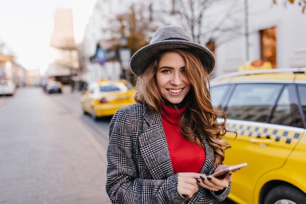 Businesslady guapa con sombrero gris sonriendo suavemente, caminando por la calle. Impresionante modelo femenino de pelo oscuro en abrigo de pie junto a un taxi en la mañana.