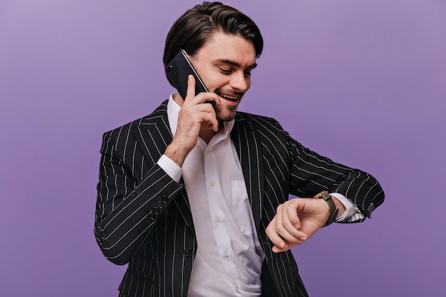 Caballero joven ocupado con camisa blanca de pelo moreno y traje de rayas negras hablando por teléfono mirando el reloj y sonriendo contra el concepto de negocio de fondo violeta