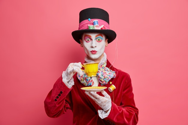 Foto gratuita el caballero sorprendido tiene la imagen del personaje del país de las maravillas viste un sombrero de guantes de encaje de traje aristocrático y bebe té tiene maquillaje de calavera colorido aislado en una pared rosa