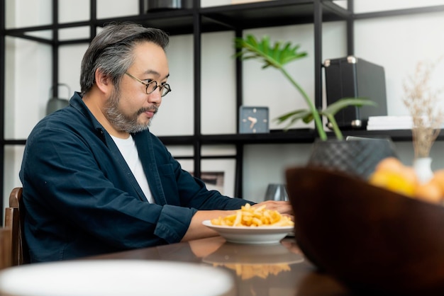 Cabello y barba plateados. Hombre de negocios independiente que trabaja en casa detrás de una computadora portátil con una comunicación súper ocupada que llama a un teléfono inteligente. Trabajo remoto y familia.