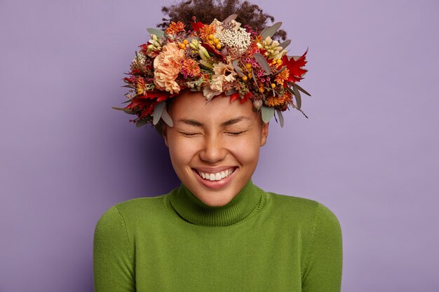 En la cabeza de la adorable mujer afro se ríe alegremente viste una corona otoñal hecha a mano, sonríe ampliamente, viste un cuello alto verde