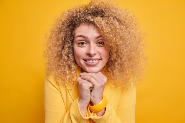 En la cabeza de la encantadora mujer de pelo rizado mantiene las manos bajo la barbilla sonríe suavemente estando de buen humor