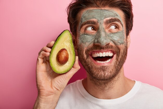 En la cabeza de un hombre moreno guapo con amplia sonrisa dentuda, sostiene una rebanada de aguacate cerca de la cara, usa una máscara de arcilla casera nutritiva para reducir los puntos oscuros, usa una camiseta blanca informal. Concepto de bienestar