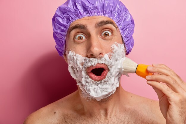 En la cabeza del hombre sorprendido usa gorro de baño, se para con el torso desnudo, se prepara para el día, se afeita la barba incipiente, se aplica crema de afeitar con un cepillo, tiene un aspecto asombrado