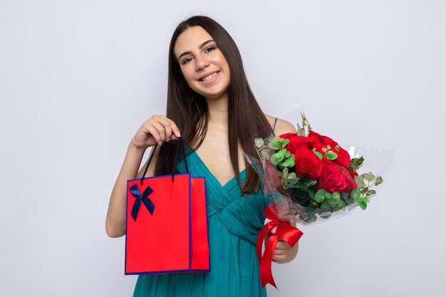 Cabeza inclinada sonriente hermosa joven en el día de la mujer feliz sosteniendo ramo con bolsa de regalo aislado en la pared blanca