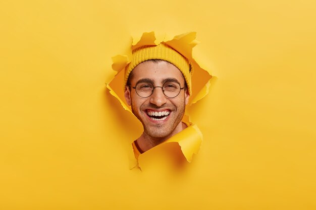 En la cabeza de un joven sin afeitar positivo sonríe ampliamente, lleva gafas ópticas redondas, casco amarillo