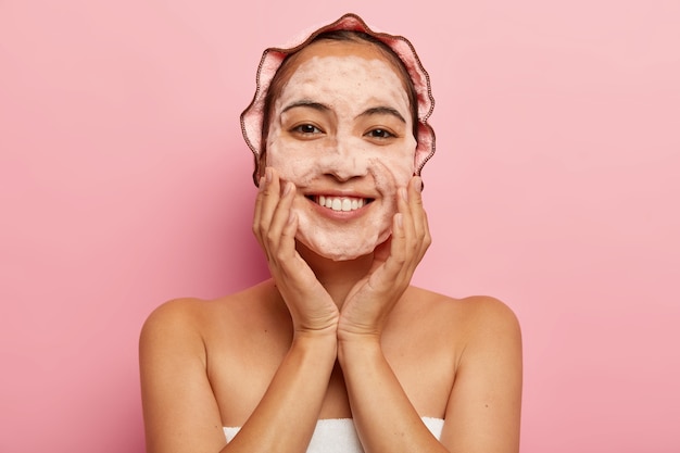 Foto gratuita en la cabeza de una joven coreana toca una piel suave e impecable, se lava la cara con jabón higiénico con limpiador espumoso, envuelta en una toalla, tiene gorro de baño en la cabeza, aislado en la pared rosa. concepto de limpieza