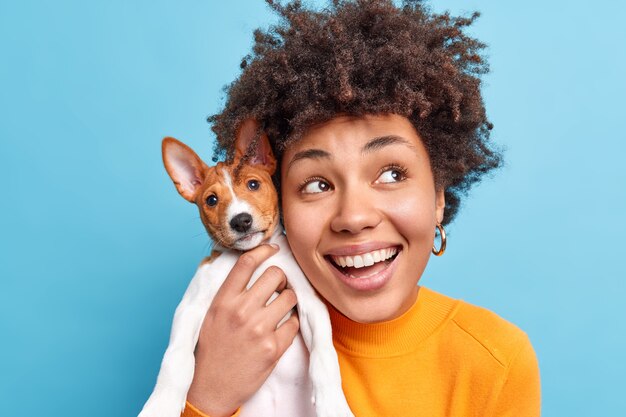 En la cabeza de la mujer afroamericana de piel oscura sonriente feliz sostiene un perro de raza agradable que expresa emociones positivas tiene una expresión soñadora que va a caminar con su mascota favorita. Concepto de personas y animales.