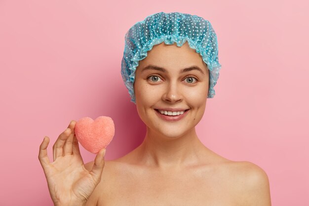 En la cabeza de una mujer europea alegre con expresión feliz, sonrisa suave, dientes perfectos, usa gorro de ducha, sostiene una pequeña esponja en forma de corazón, se ducha, tiene la piel limpia y saludable. Concepto de higiene