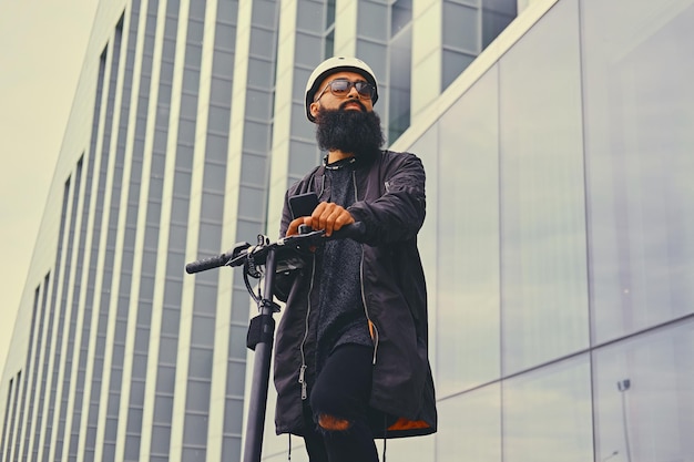 Cabeza rapada tatuada, hombre barbudo usando un teléfono inteligente sobre el fondo de un edificio moderno después del paseo en scooter eléctrico.