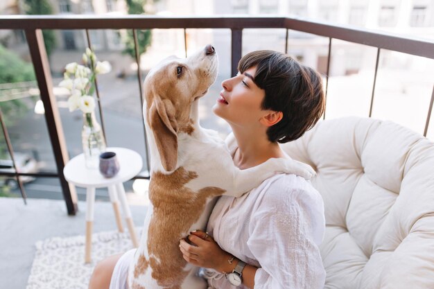 Cachorro beagle divertido mirando hacia arriba sentado en las rodillas de la niña bonita en la mañana. Retrato de mujer morena inspirada posando sobre fondo borroso con expresión de cara tranquila con mascota.